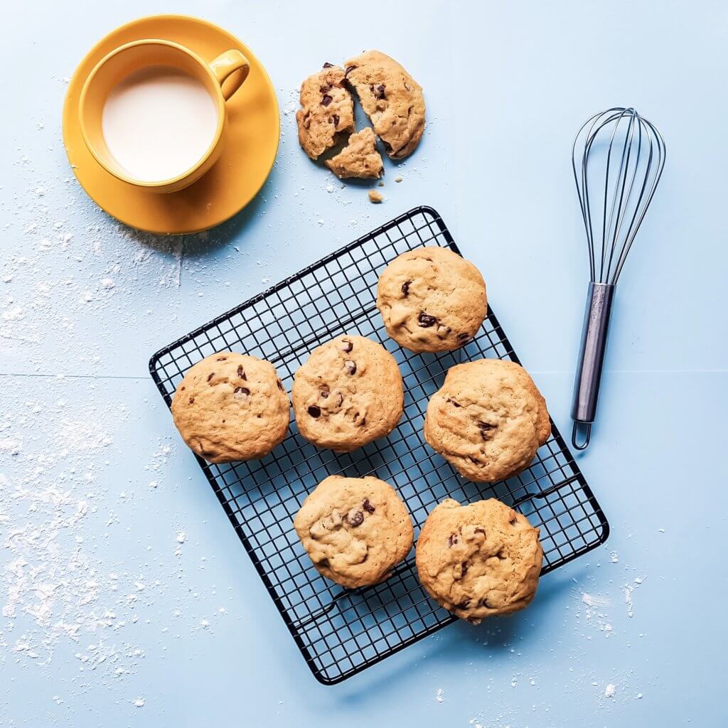 An image of 6 chocolate chip cookies from above sitting on a drying rack. Flour is sprinkled around, as if they were just made. Nearby is a whisk, and a yellow saucer and teacup filled with milk with a broken cookie next to it.