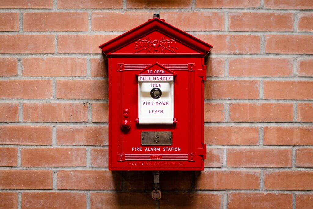 An old-fashioned school fire alarm box is on a brick wall.