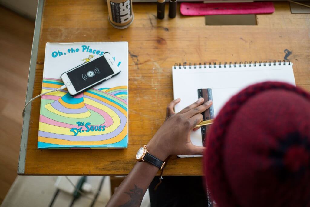 a person with a maroon felt hat and dark skin holding a ruler and pencil over a sketchbook that is on a scuffed wooden work table. To their left of their sketchbook is “Oh The Places We’ll Go” by Dr. Seuss. A phone is on top of the book that has the audio play button displayed on it.